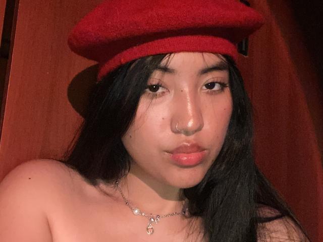 Elizaa, com longos cabelos pretos, usando um chapéu vermelho, um piercing no nariz e um colar delicado, tem lábios rosados e brilhantes e um ombro descoberto, olhando diretamente para a câmera..