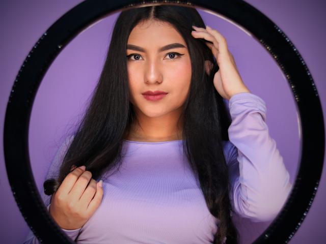 AnieDemond with long black hair, wearing a purple long-sleeve shirt, looking at camera, hand in hair, through a circular black frame, showing her cute, sexy face.