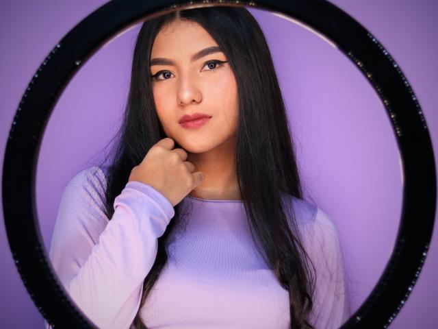 AnieDemond has long black hair, a cute face, and a hand near her neck. She's wearing a light purple top, looking at the camera with subtle makeup.