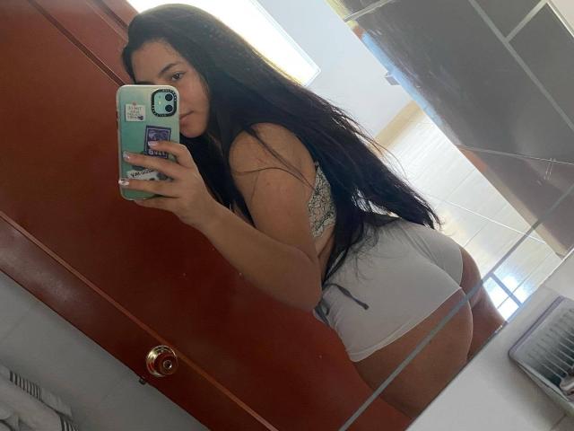 AnieDemond with long black hair, wearing a grey bra and white booty shorts, taking a mirror selfie, showing her big ass and tattoo on her back, holding a green phone.