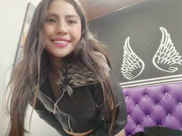 PaulaCeleste with long dark hair, smiling, wearing a black zip-up jacket with fur trim, purple-tufted headboard in background. Her pink lipstick and black top are visible.