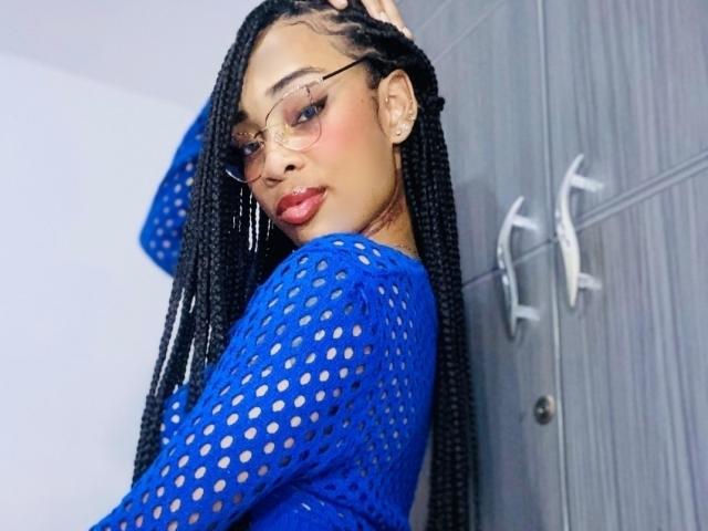 ElaHott with long black braids, wearing a blue mesh top, glasses, and red lipstick, standing against a gray door, looking back over her shoulder with a sultry expression.