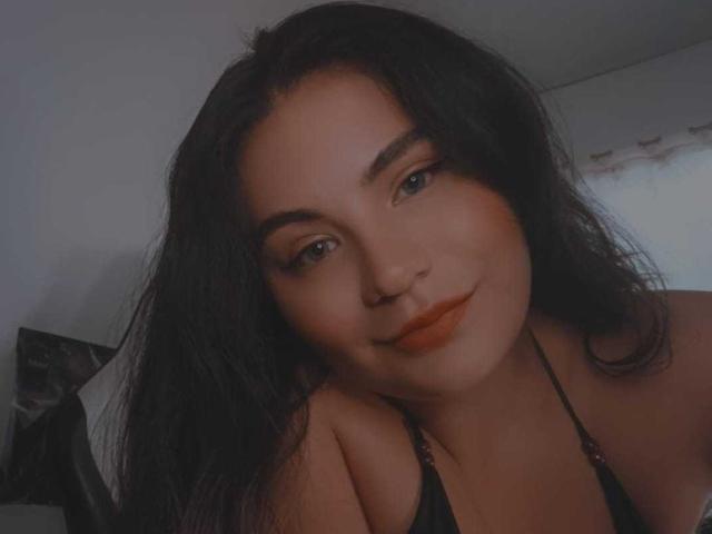 GigiQueen has dark, wavy hair and orange lipstick. She's smiling softly, with a subtle, sexy look, wearing a black halter top, showing her tits.