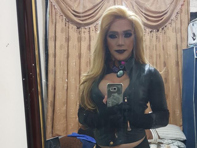 QueenOfPainSasha with long blonde hair, black lipstick, and a leather top, holding a phone in front of a mirror, showing her tits. Wearing black gloves, necklace with purple stones.