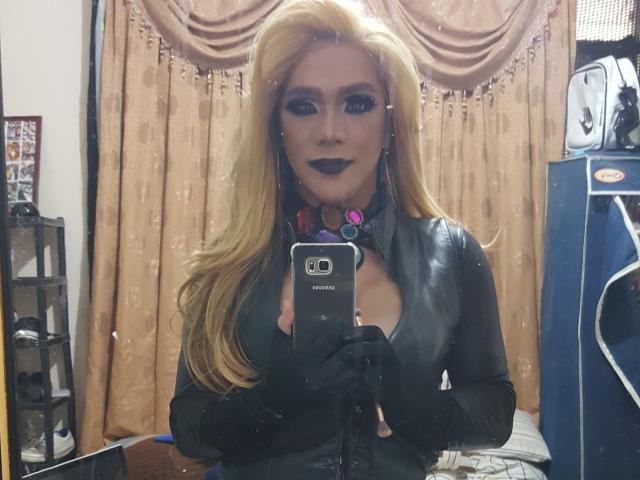 QueenOfPainSasha with blonde hair, dark lipstick, and black leather outfit, holding a phone, wearing black gloves, and a colorful choker, taking a mirror selfie. Boobs visible.