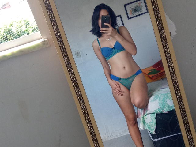 LuaMendes in blue bra and green panties, mirror selfie, holding phone, short black hair, medium tits, slim, standing with one leg up, room with bed and window.
