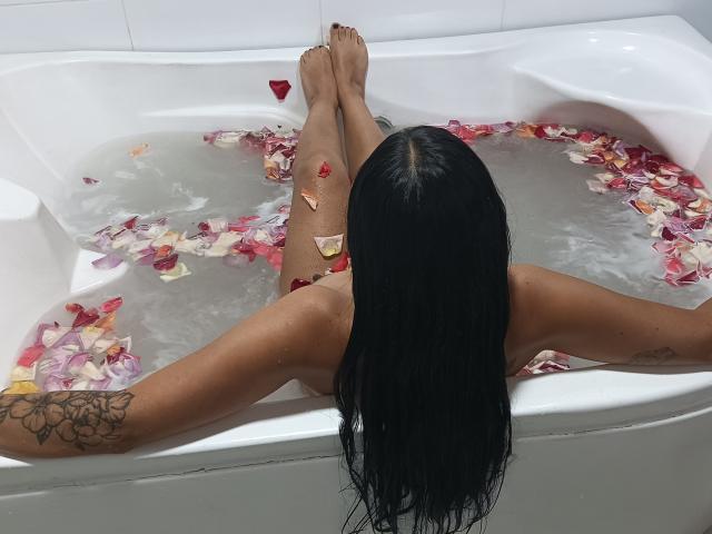 GeshaLover with long black hair, arms spread, legs up in white bathtub filled with water and red rose petals, tattoos on arms, back to camera.