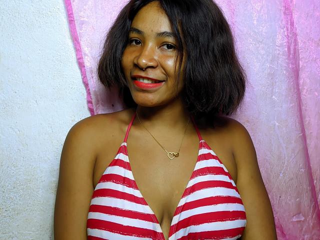 KonyHot with brown skin and red lipstick, wearing a red and white striped bikini top, smiling, has small tits, and a heart necklace against a pink and white backdrop.