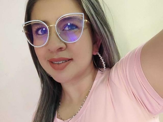 MelindaBoobs with purple-eyed glasses, pink shirt, and gray hair, wearing pink earrings and a silver necklace, smiling with metal teeth.
