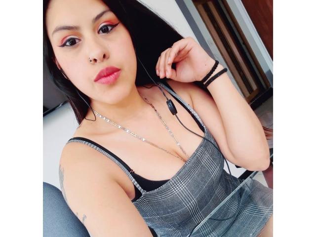 ElaiaaBakerr with black hair, nose ring, and pink lipstick, wearing a checkered top, exposing her black bra and small tits, holding earbuds, and a tattoo on her arm.