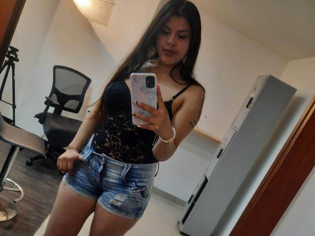ElaiaaBakerr with long brown hair, black lace top, and denim shorts, holding a phone, showing her small tits and cute ass, standing in a room.