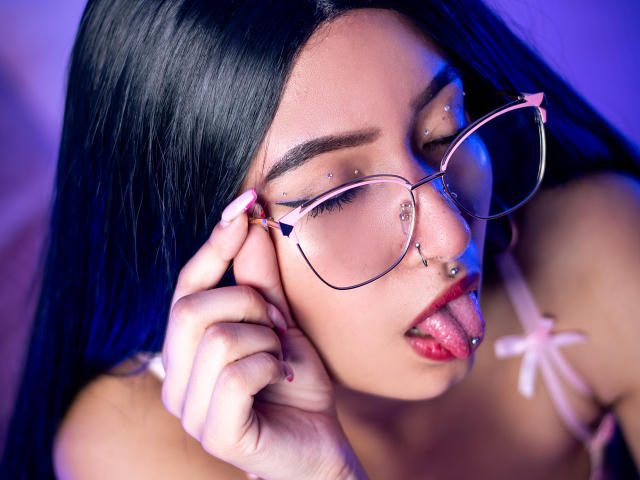 SophiaAndTony with black hair, nose piercings, and glasses, sticking out pink tongue, eyes closed, wearing pink straps, blue light, and piercings on eyebrows and lip.