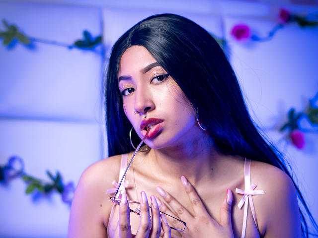 SophiaAndTony with long black hair, pink lipstick, and hoop earrings, sucking on glasses while touching her tits with pink-manicured nails, under blue lighting.