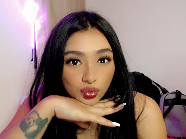 AndreaPretty with long black hair, pouty red lips, and a tattoo on her right hand, blowing a kiss, looking directly at the camera. She has thick eyelashes and white nails.