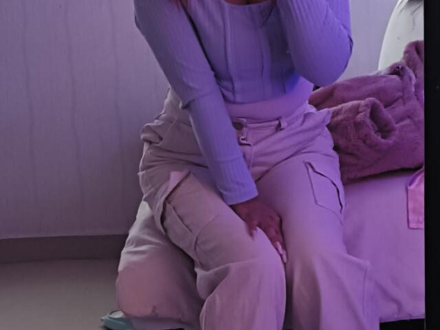 ShofiaWett sentada en una cama, vistiendo una camiseta de manga larga azul claro con textura acanalada y pantalones cargo blancos, con las manos sobre su muslo, en una iluminación púrpura tenue..