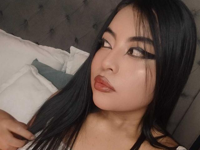 AlexaMadisson has long black hair, thick black eyebrows, and glossy brown lips. She's looking to the side with a slight pout, wearing a white top.