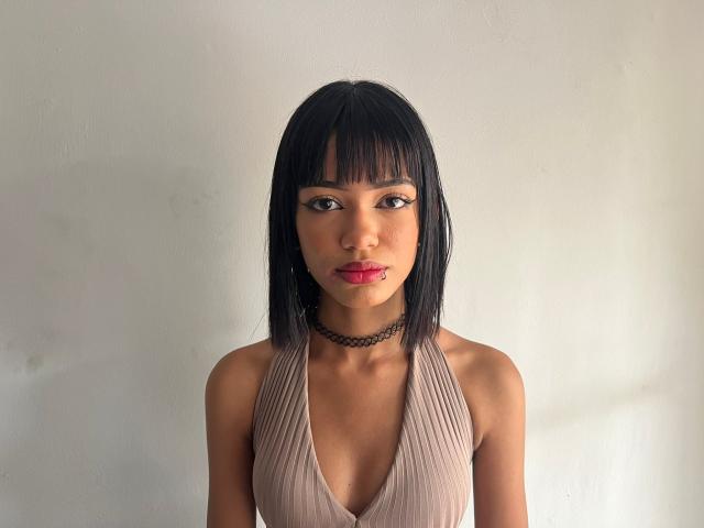 KylieGreenX has black bob hair, brown eyes, pink lipstick, choker, and a deep V-neck beige top showing small tits. She's staring forward with a serious expression.