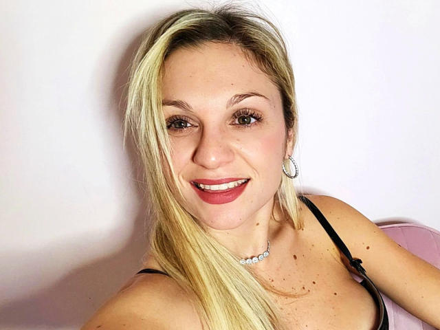 GeileVivienne-hot has blonde hair, red lipstick, and is smiling at the camera. She's wearing a black top, pearl necklace, and silver earrings, showing her tits.