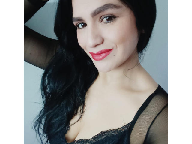 CameronViviane with black hair, red lipstick, and black lace top, showing her tits, smiling, arm raised, looking at camera.