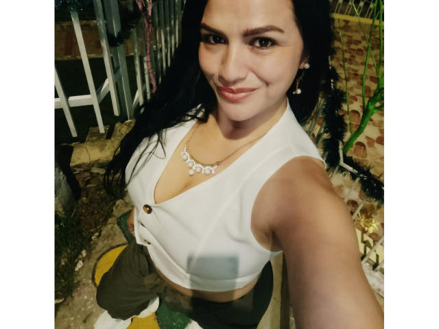 CameronViviane, with long black hair, smiling, wearing a white sleeveless top showing her tits, green pants, and a necklace, taking a selfie outside at night.