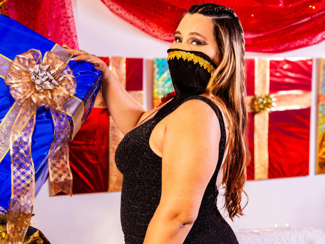 SalmaAzaman with long brown hair, wearing a black glitter dress and gold-trimmed black mask, holding a blue gift with gold ribbon, showing her tits and looking at camera.
