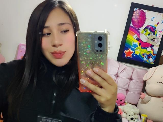 ValeThomson with long black hair, wearing a black zip-up, holding a glittery phone, pouting pink lips, pink blush, cute, taking a selfie, pink bed, colorful wall art.