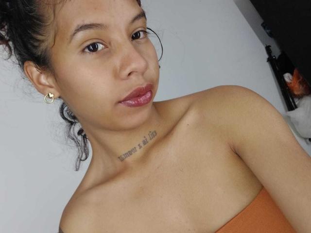 VelvetSirens has a cute face, medium brown skin, and small tits, with a tattoo on her neck and wearing a strapless orange top, and her lips are shiny.