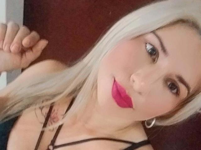 ChristinaSilva with long blonde hair, pink lipstick, hoop earrings, and a black strappy top, showing a cute smile and raised hand. Boobs visible under top.