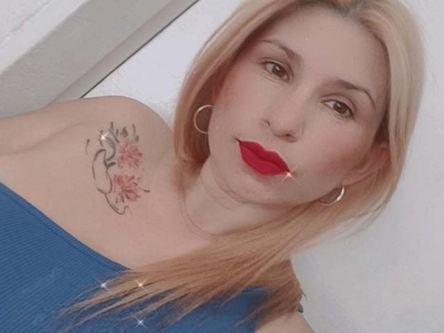 ChristinaSilva with blonde hair, red lipstick, hoop earrings, blue off-shoulder top, and a tattoo of flowers on her right shoulder, looking at the camera.