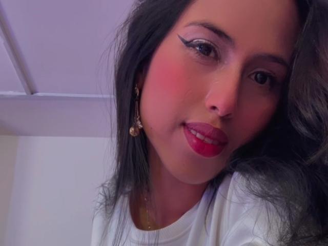 MyaaTyler, con rossetto rosso, eyeliner nero e fard rosa, indossa una camicia bianca, ha lunghi capelli neri e orecchini dorati, e sorride leggermente. Le sue natiche non sono visibili..