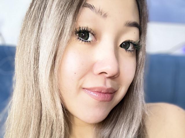 RenyLime has light brown skin, long blonde hair, green eyes with long lashes, and glossy pink lips. Her nose is small and she's smiling slightly.