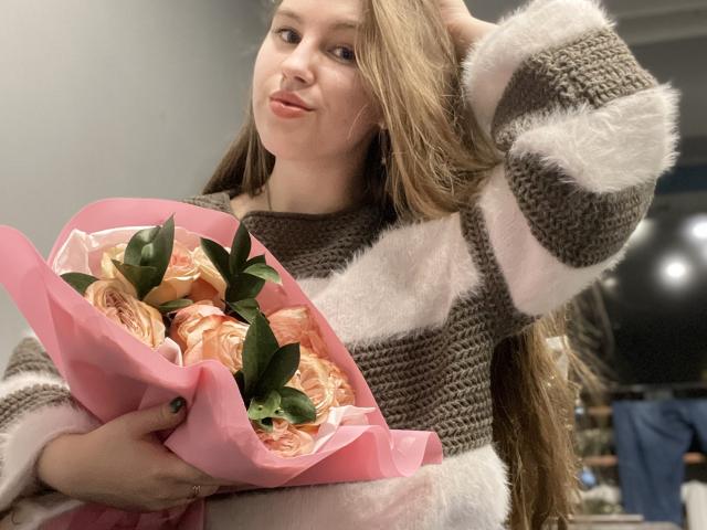 Dooris with long brown hair, pouty lips, wearing a fuzzy brown and white striped sweater, holding pink-wrapped roses in her hand, looking at camera with a slight smile.