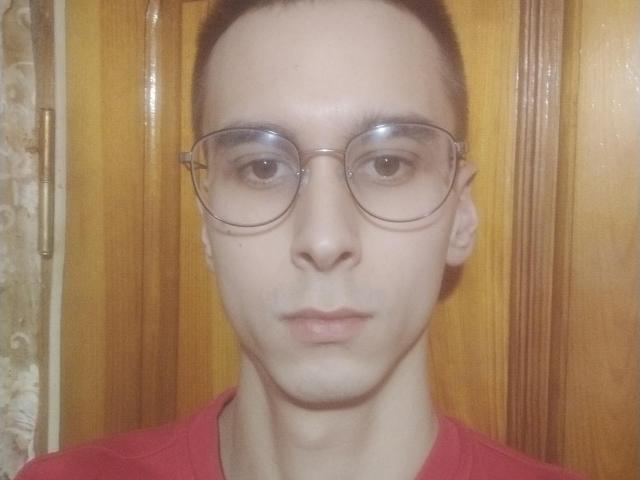 WinterBrian, con cabello corto y castaño, que usa gafas redondas transparentes y una camisa roja, tiene un tono de piel pálido y una expresión neutra, y está de pie frente a una puerta de madera..