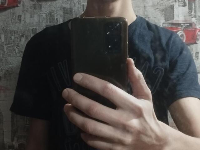 WinterBrian, la main tenant un téléphone noir, cachant son visage, portant une chemise bleu foncé, avec un objectif de caméra visible sur le téléphone, dans un environnement sombre et peu attrayant..