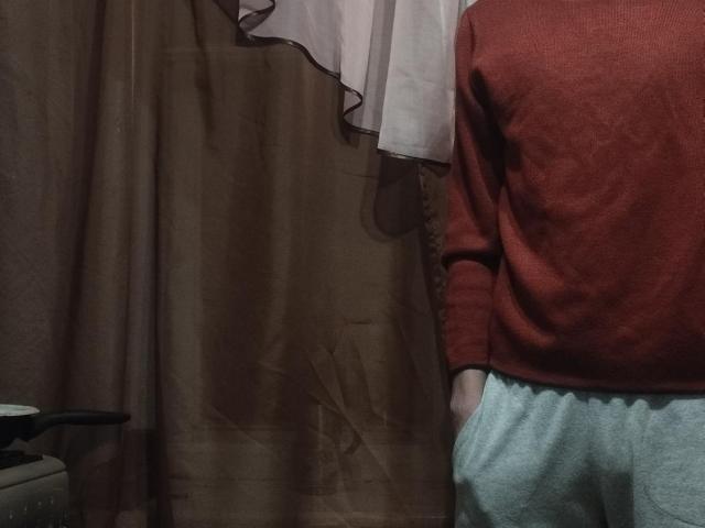 WinterBrian steht vor braunen Vorhängen, trägt einen roten Pullover und hellgraue Hosen, wobei Gesäß und Hände sichtbar sind, und ist leicht zur Seite gedreht..