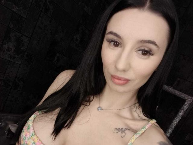 BriannaBratty with black hair, brown eyes, and pink lipstick, wearing a floral top, showing small tits and tattoos on her chest, looking at the camera with a slight smile.