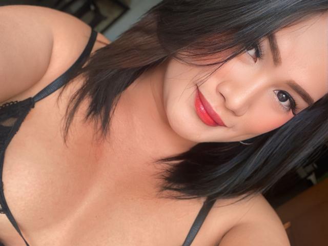 AlwinaForU has black hair, red lipstick, and black lingerie, showing her tits and a cute smile. Her skin is tan, and she's looking at the camera.