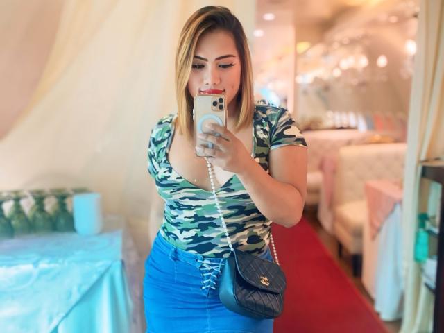 AlwinaForU with blonde hair, camo top, and blue skirt, holding phone, looking down, wearing black purse, standing, red carpet, blue table, beige room, smiling, sexy.
