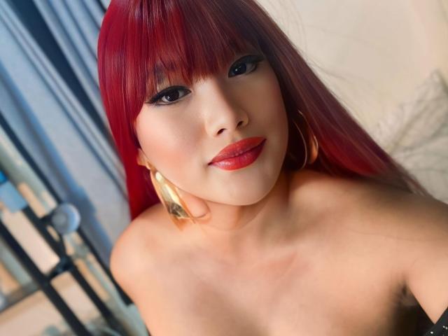 AlwinaForU with red hair, red lipstick, and gold hoop earrings, showing her bare shoulders and smiling at the camera. Her tits are partially visible.