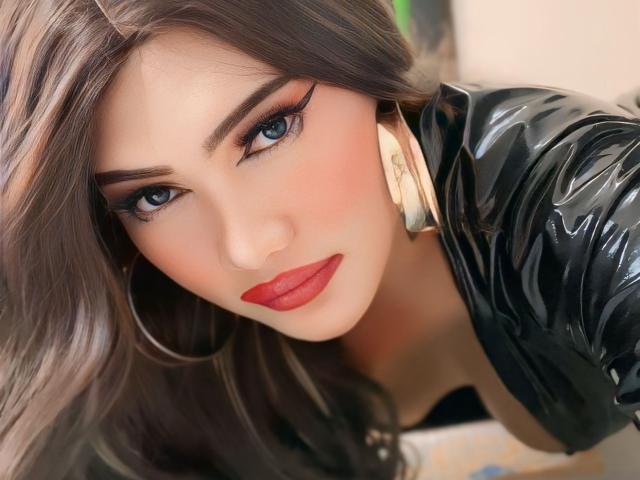 AlwinaForU with glossy black latex, red lipstick, blue eyes, and long brown hair, looking seductively at the camera, wearing gold hoop earrings. Her ass is partially visible.