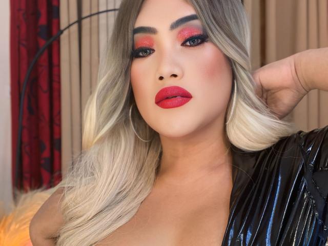 AlwinaForU has blonde hair, red lipstick, and heavy eye makeup. She's wearing a black, shiny top, with her hand behind her head. Her tits are slightly visible.
