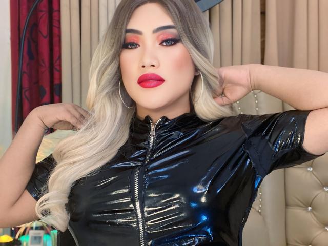 AlwinaForU with long blonde hair, red lipstick, and heavy makeup, wearing a shiny black latex top, hands in her hair, showing off her tits, in a sexy, confident pose.