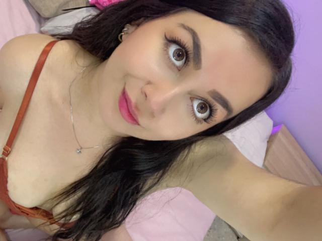 KimKolly with wide brown eyes, pink lipstick, and long black hair, wearing a brown bra, smiling slightly, holding the camera up, visible armpit, and a necklace.