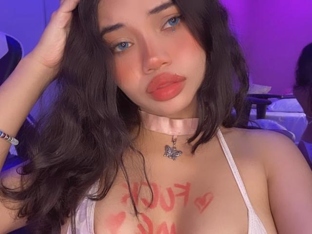 LiliMays with long dark hair, blue eyes, pink choker, and red lipstick, wearing a white top, has "You're so" in red on her chest, and a butterfly necklace.