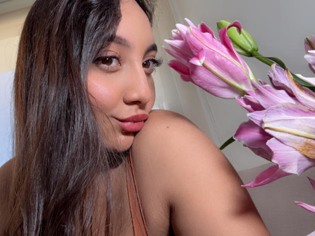 DianaFernandezz with long brown hair, subtle makeup, and pink lipstick, looking over her shoulder at pink lilies. Her shoulder is visible, showing her tan skin.