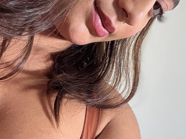 DianaFernandezz's face with shiny brown hair, pink glossy lips, and a tan skin tone, wearing a pink strap, close-up shot showing her upper chest and left side.