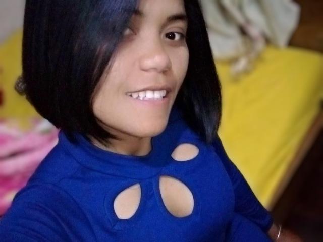ElisabethS has short black hair, smiling with white teeth, wearing a blue shirt with cutout holes showing her tits, in a selfie.
