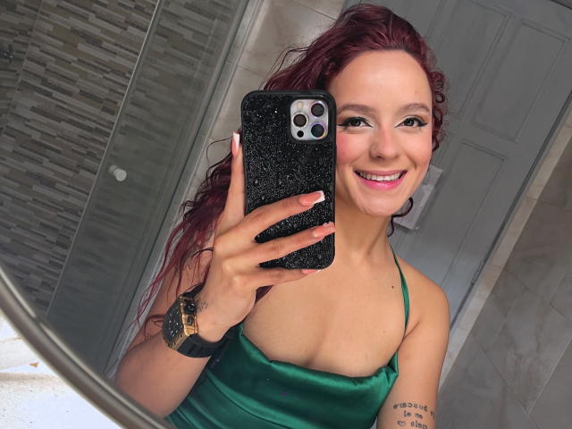 MaiaPark with red hair, smiling, wearing a green satin top, showing her tits, holding a black glitter phone, wearing a gold watch, in a bathroom mirror selfie.