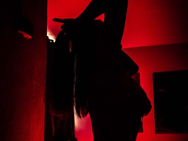 RosalindaCortez im Schatten, mit langem Haar, Armen erhoben, in einem rot beleuchteten Raum, Hände in der Nähe des Kopfes, eine schattenhafte Figur, geheimnisvoll, sexy, dunkler roter Hintergrund, Wandleuchte links..