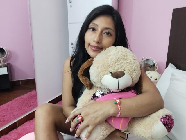 LucianaHernandez with long black hair, holding a fluffy beige teddy bear, pink ring, and pink bracelet, sitting on a bed, smiling softly, in a pink room.